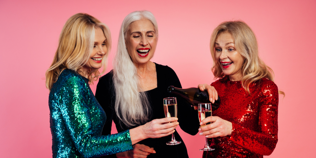 Two women hold champagne glasses while the centre pours the bottle, all laughing